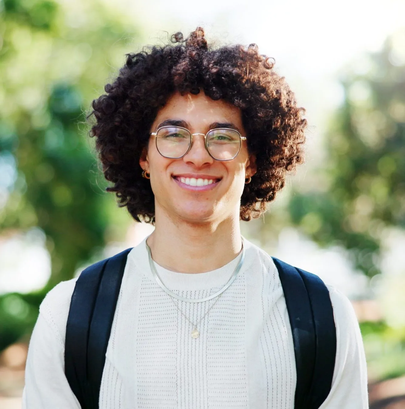Smiling person with curly hair and glasses wearing dark vest over light sweater outdoors
