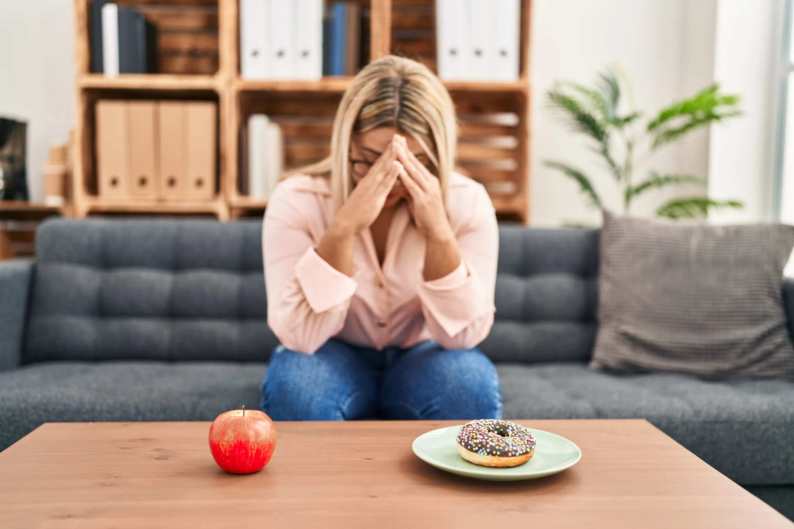 Woman sitting on a couch with her face in her hands, looking distressed, with a red apple and a frosted donut on a table in front of her