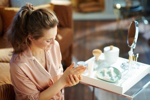 Woman applying skincare product to her hand at a vanity with cosmetic bottles and a mirror