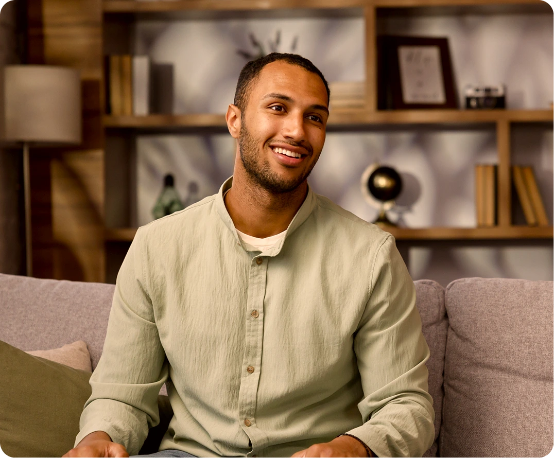 Man in beige shirt smiling while seated on a gray couch in a modern office with shelving