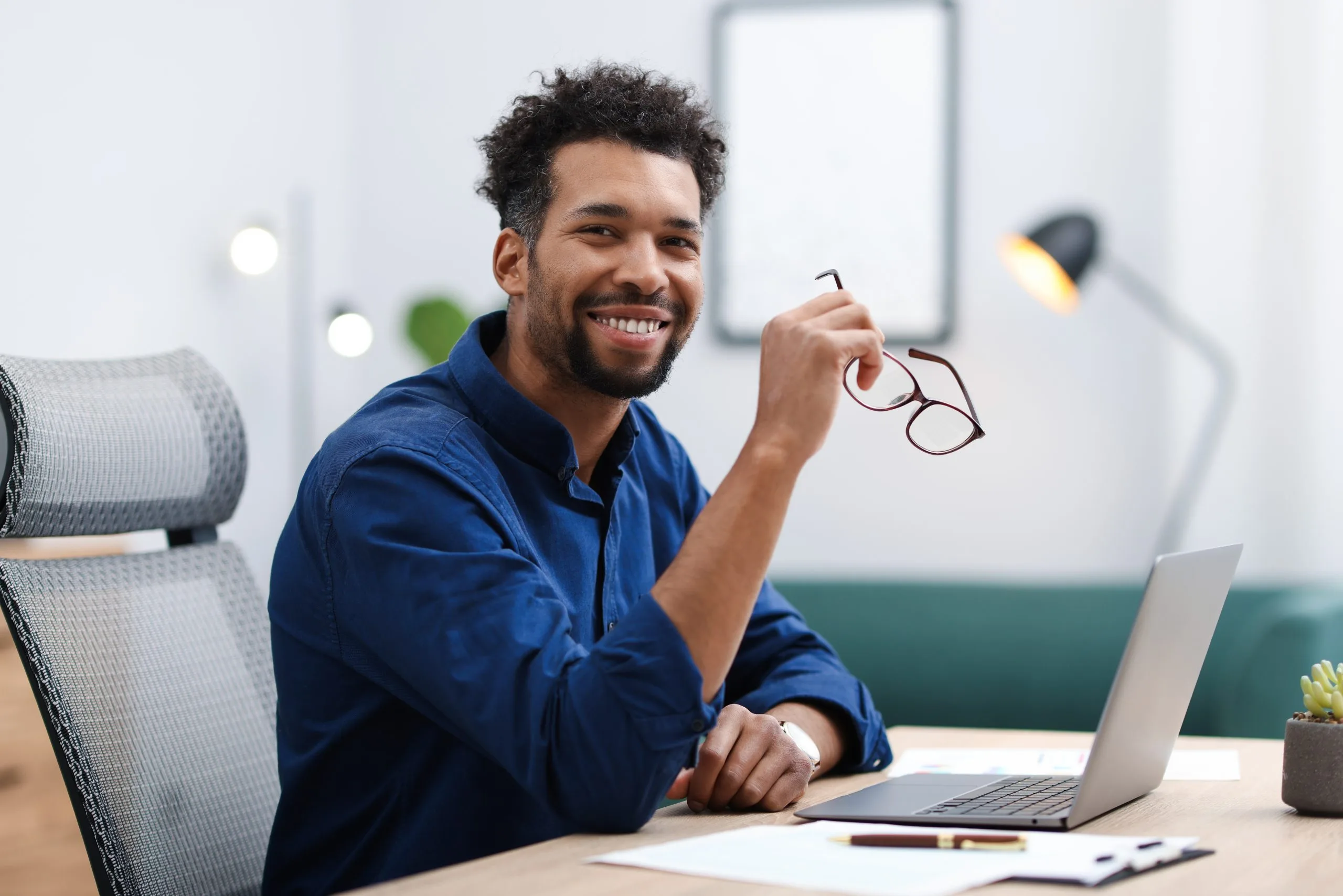 Man in blue shirt smiling at desk with laptop and glasses in modern office