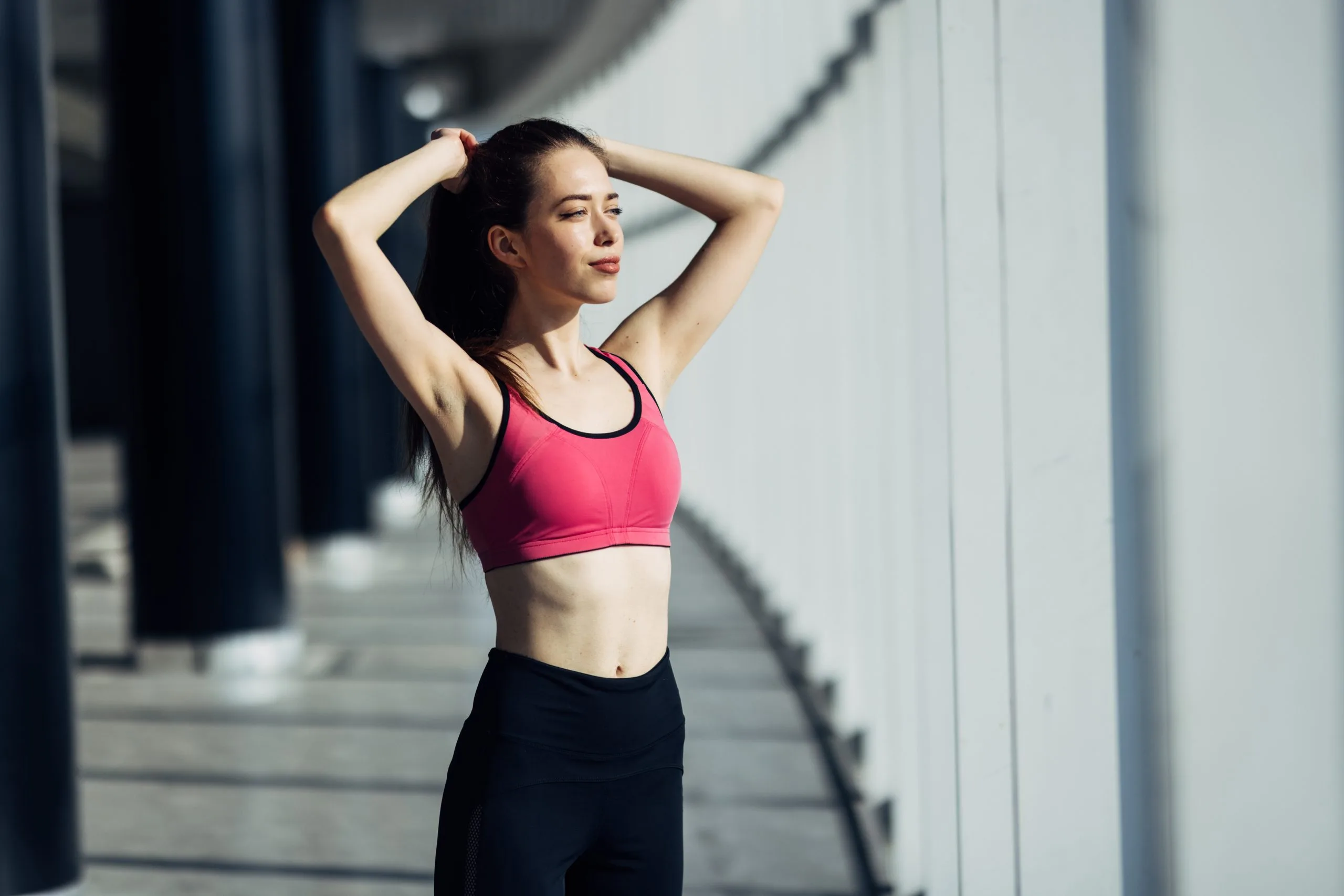 Athletic woman in pink sports bra stretching arms behind head in bright studio setting