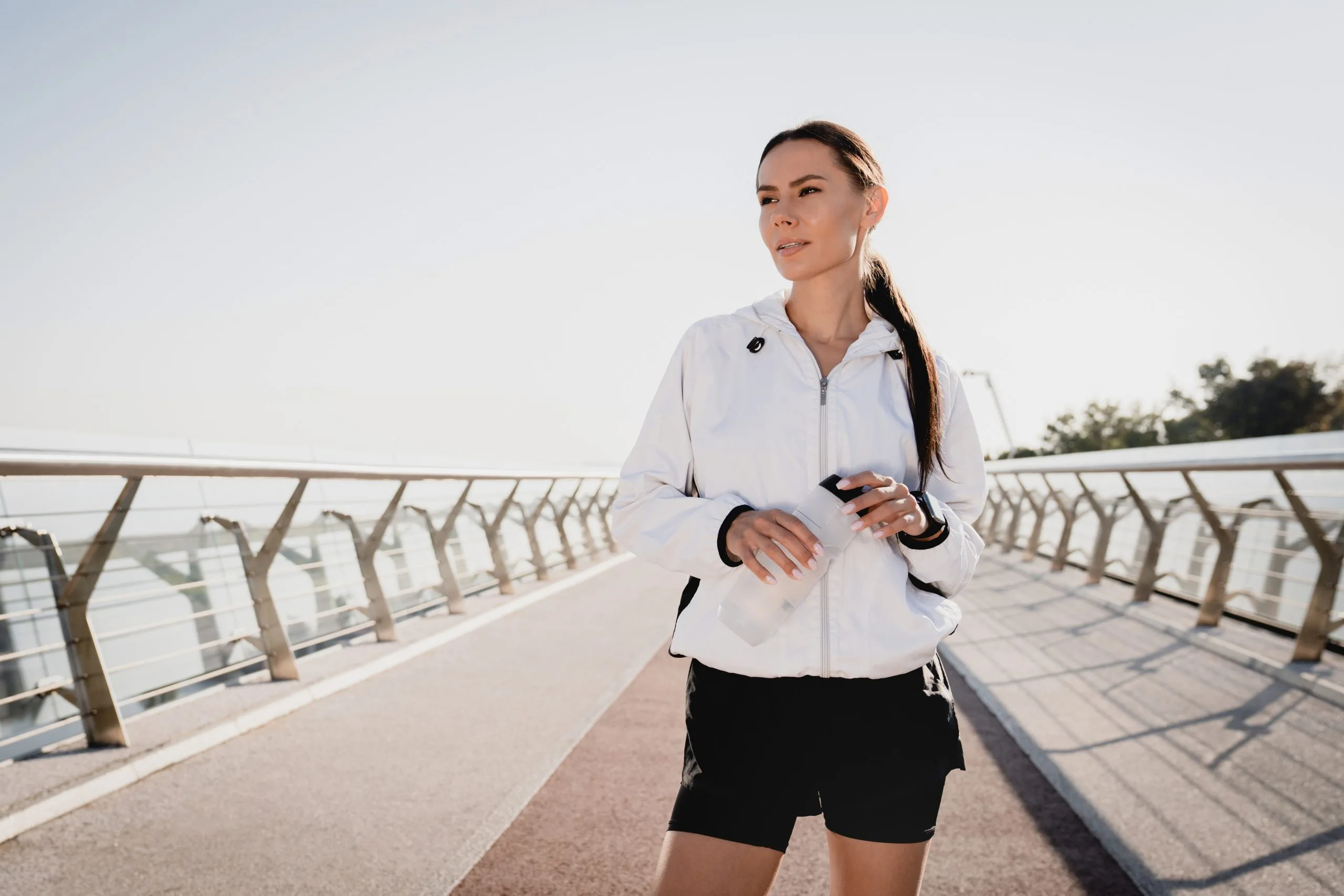 Woman in white jacket jogging on a bridge while holding a water bottle