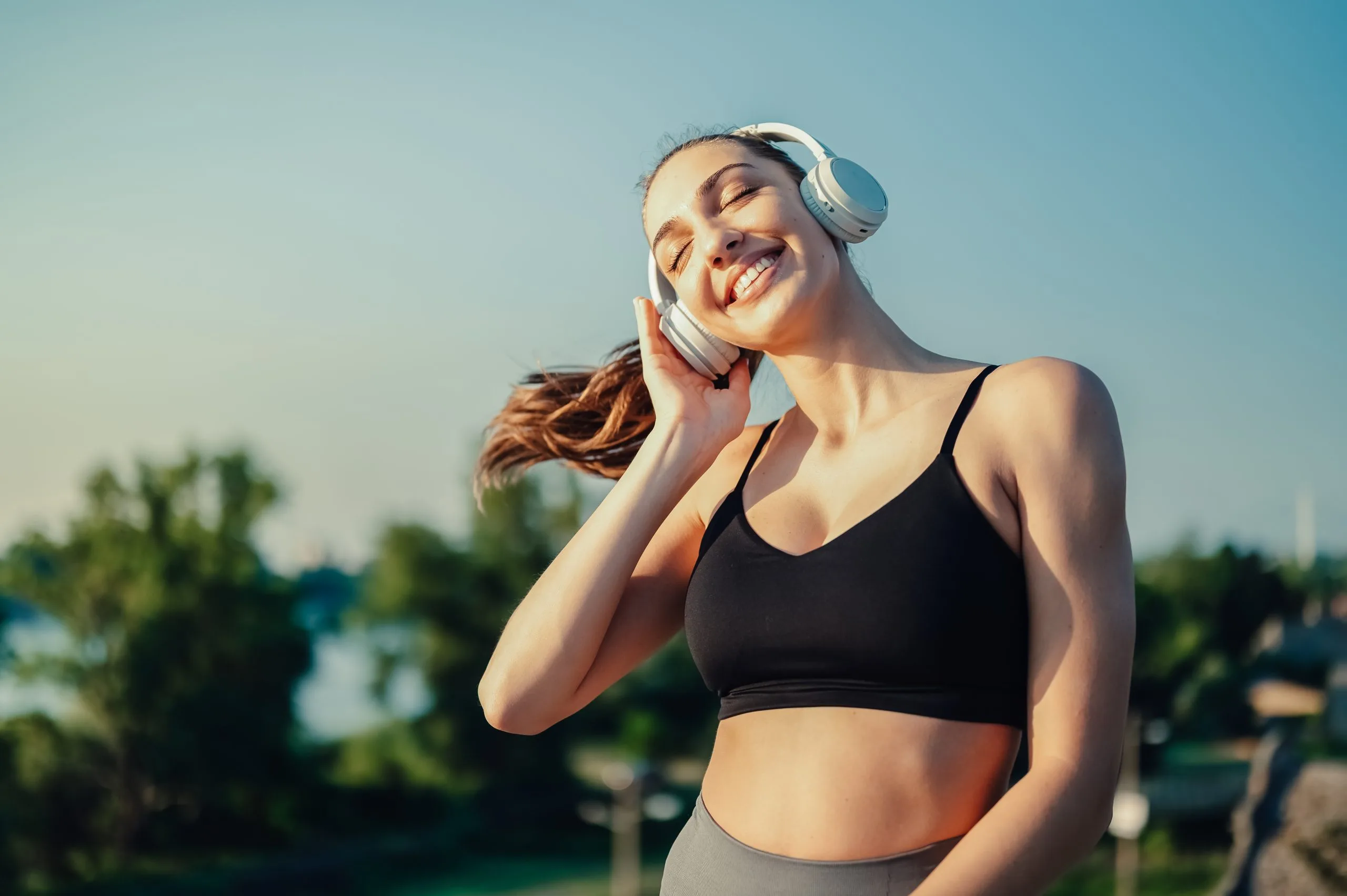 Woman in athletic wear smiling with eyes closed, wearing white headphones outdoors
