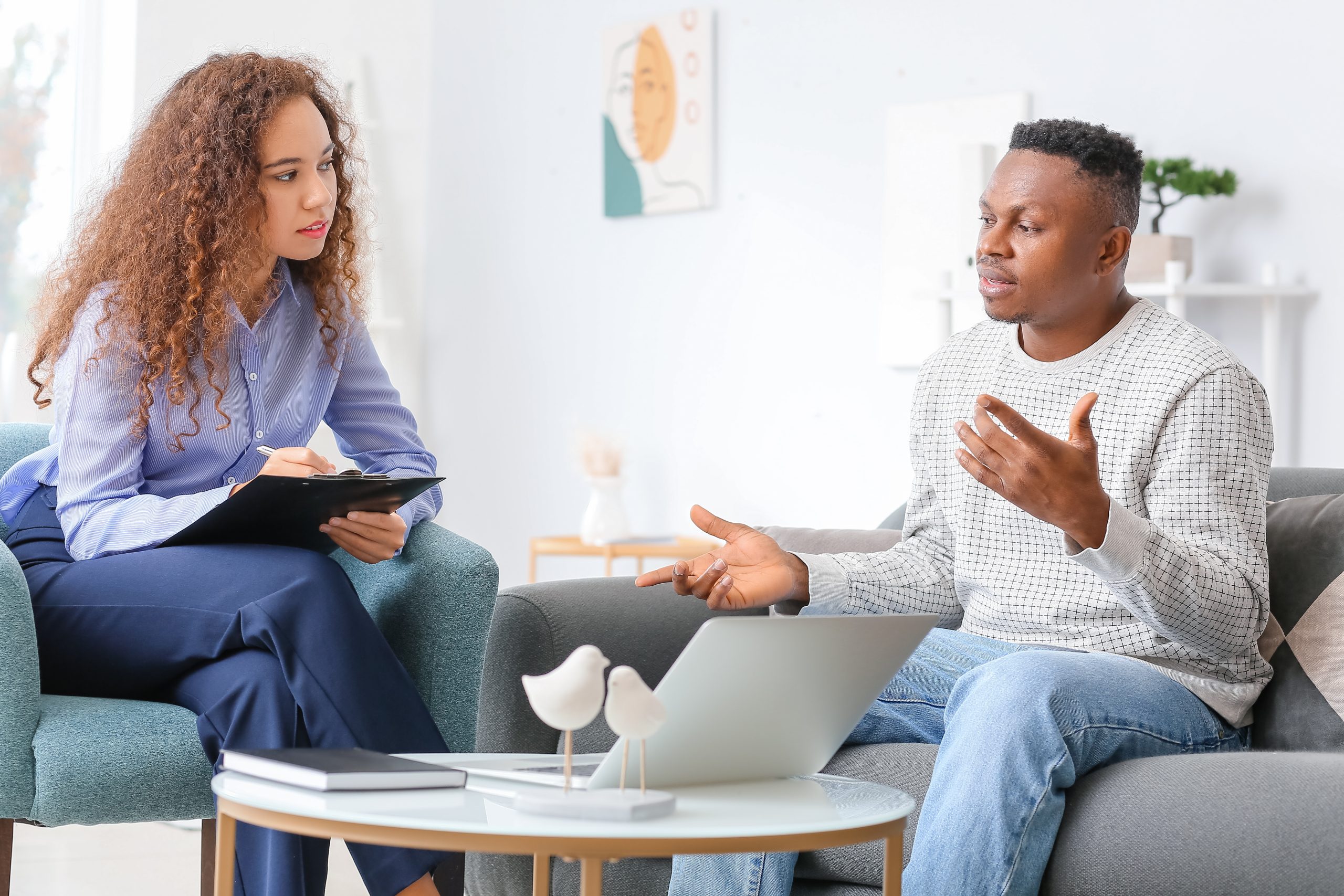 Female therapist taking notes while male client discusses on laptop during session