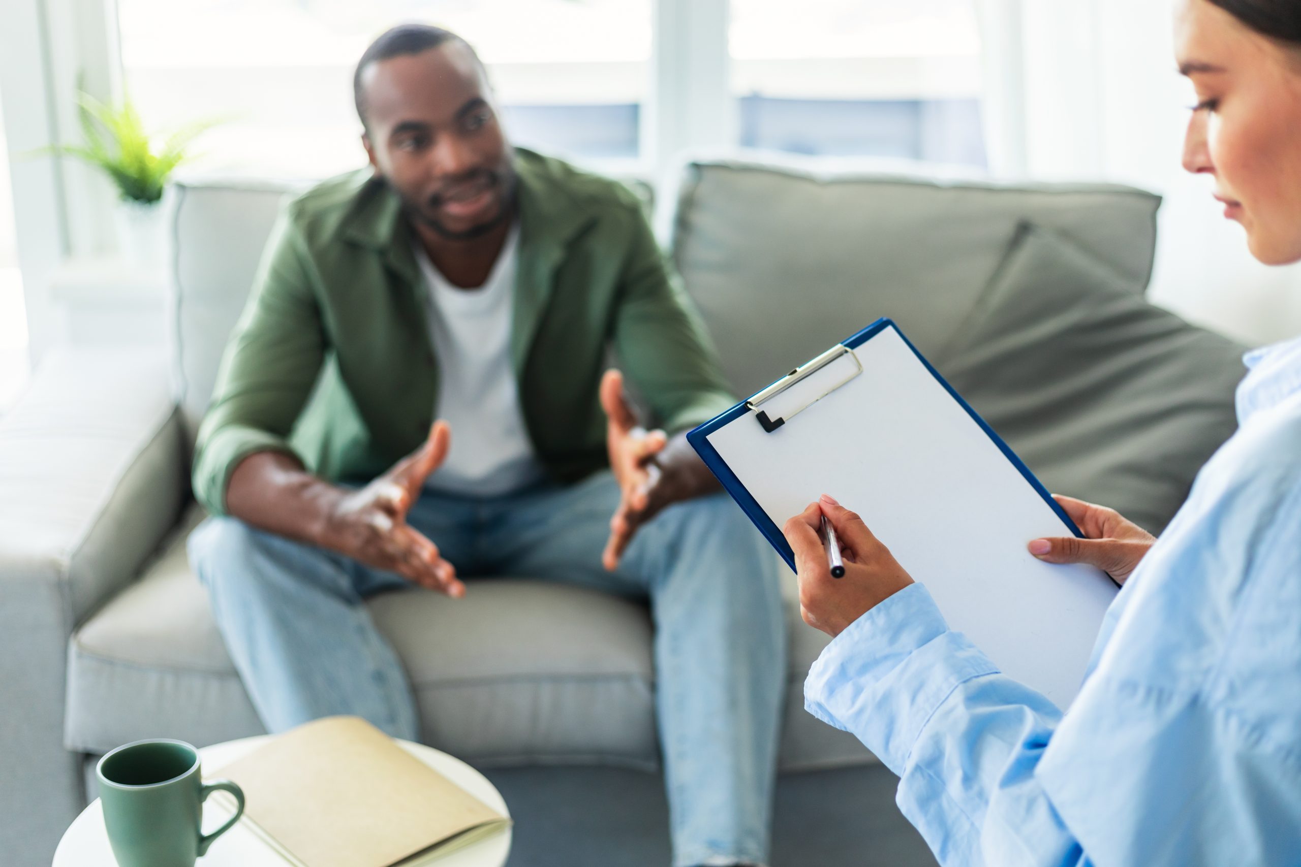 Healthcare provider holding clipboard listening to patient sitting on couch during consultation