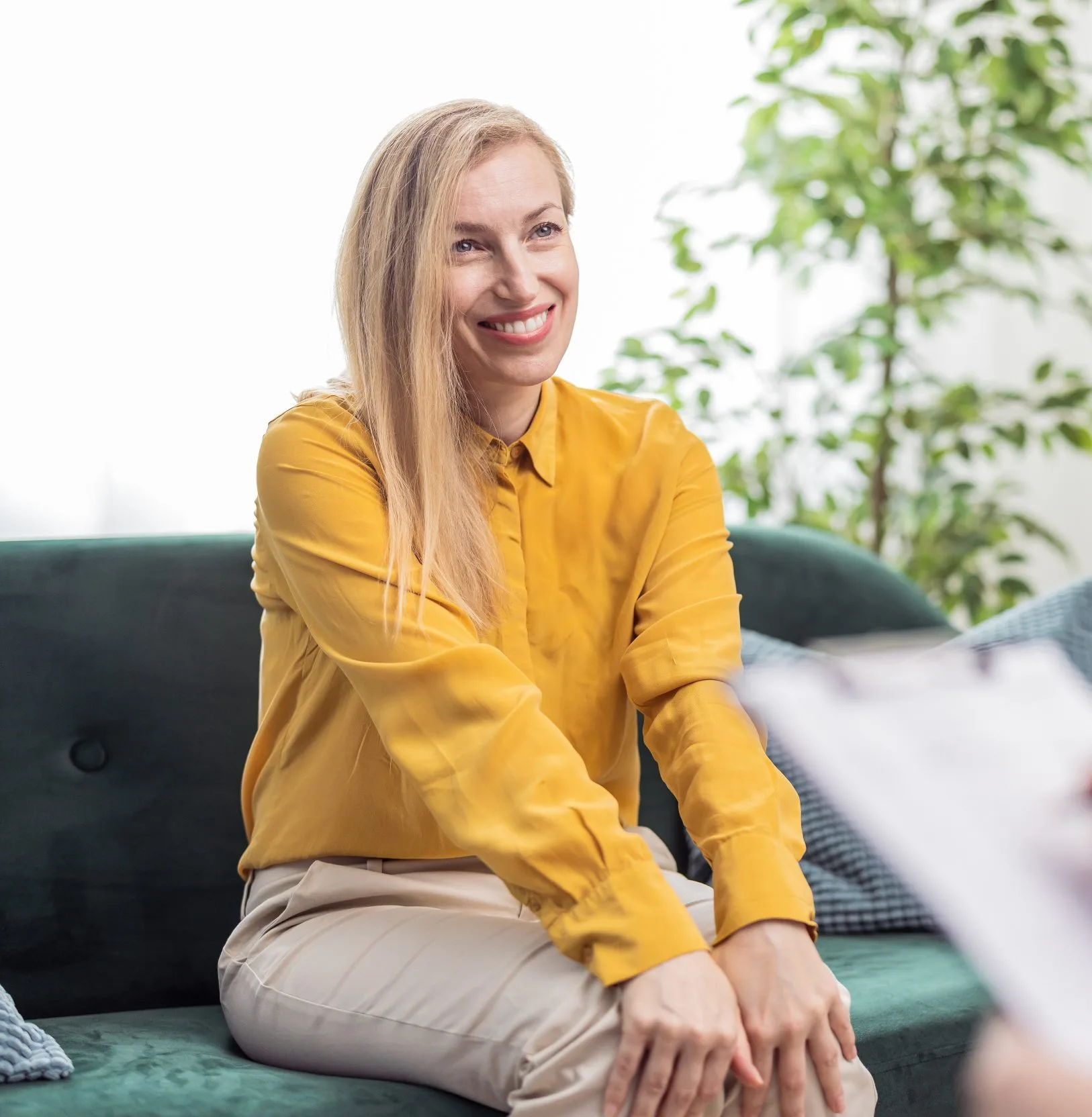 Woman in yellow shirt smiling while sitting on green chair with potted plant in background