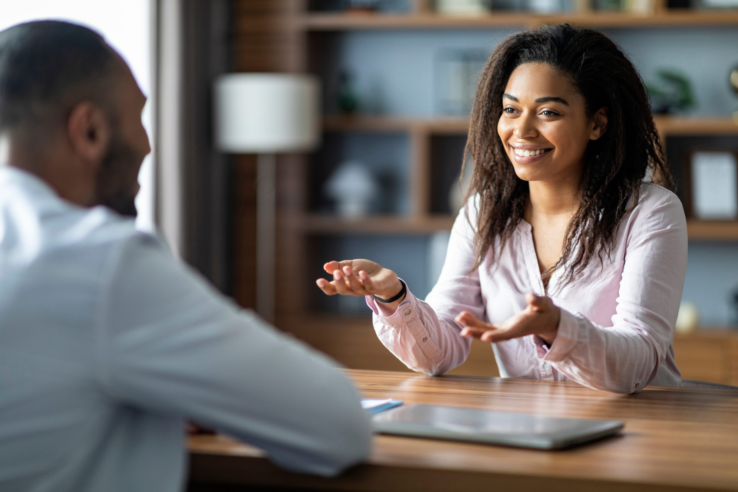 Woman in pink shirt gesturing while speaking with a man across a wooden table in an office setting