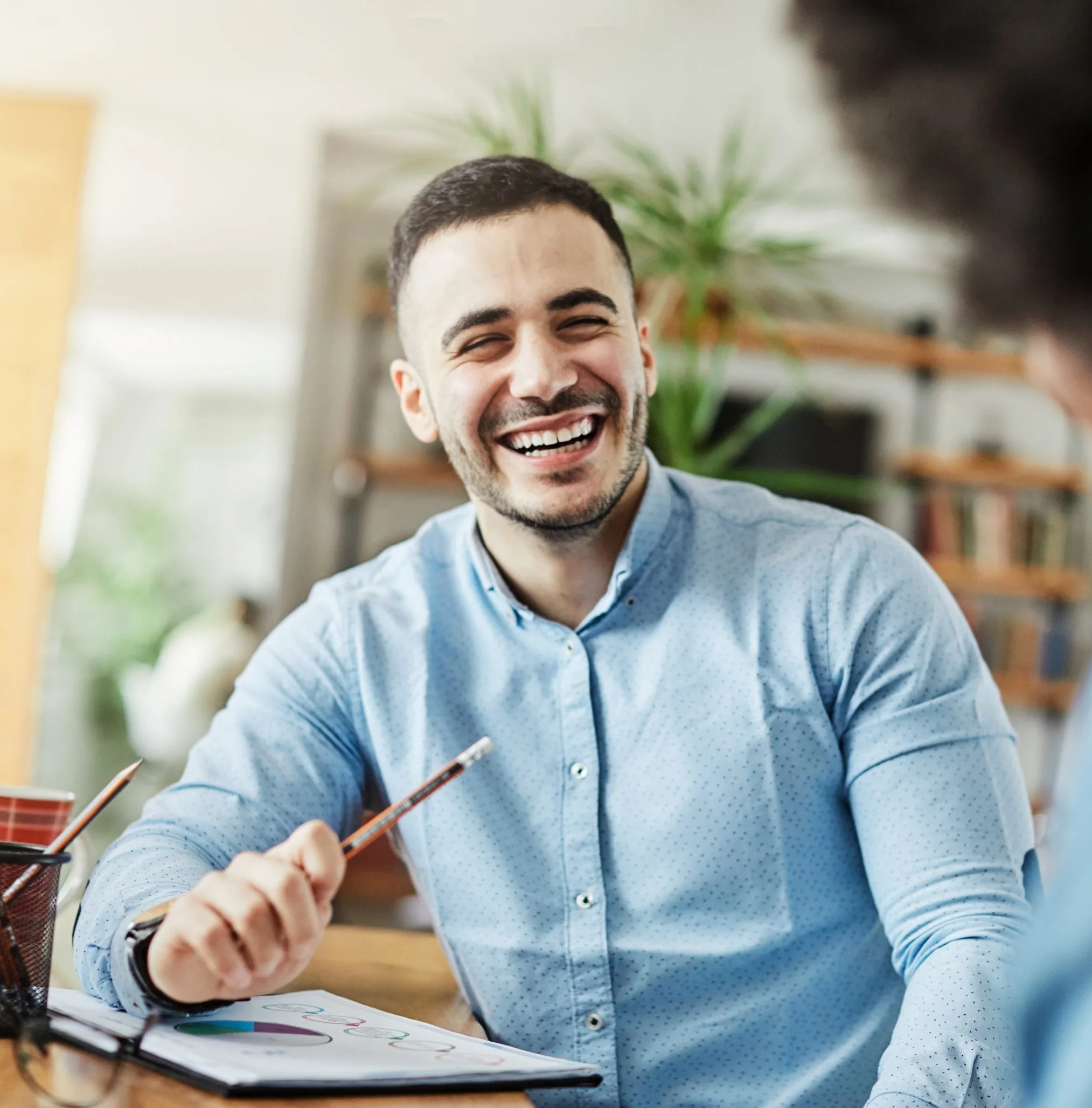 Man in blue shirt smiling while holding a pen over paperwork at a desk with plants in background