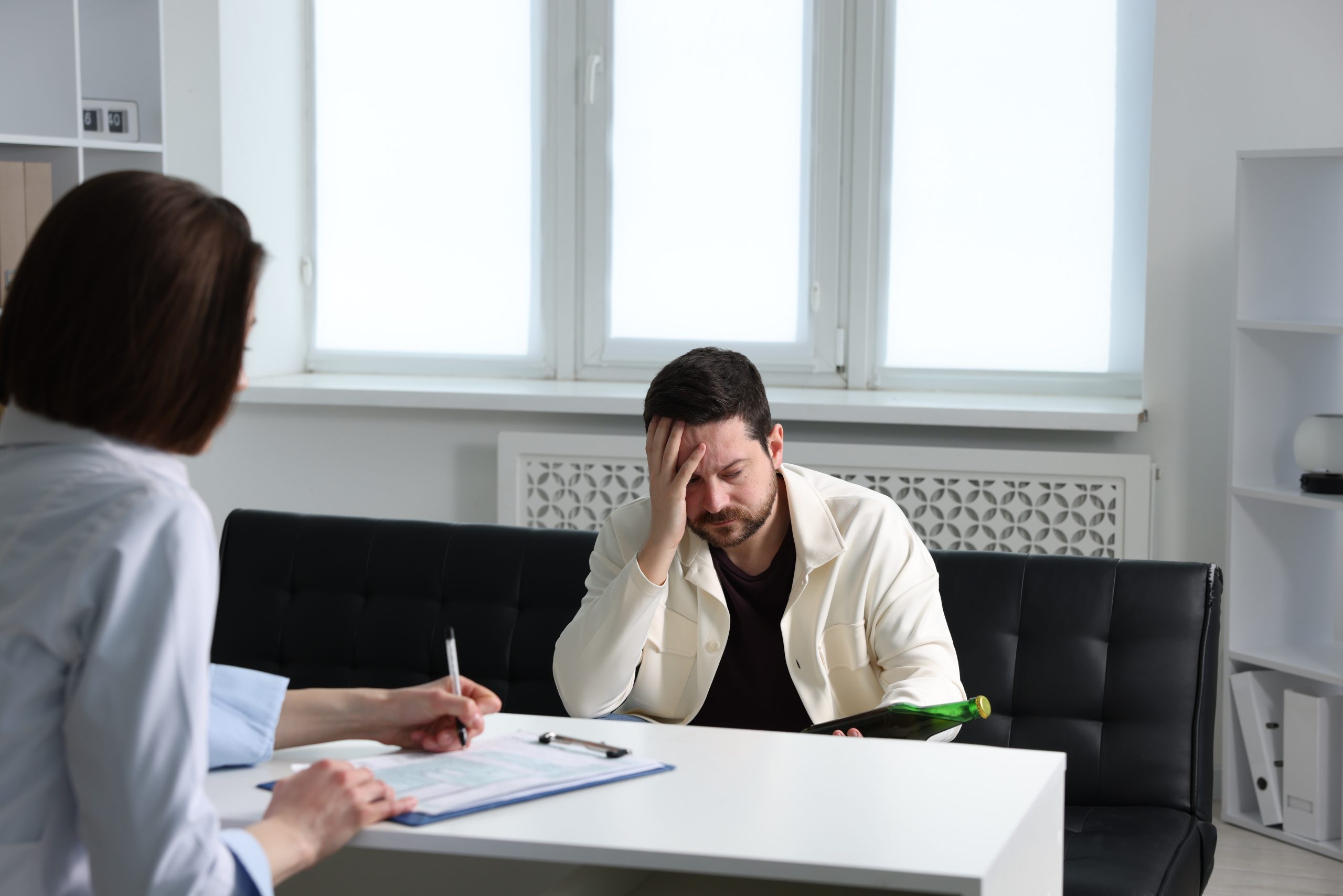 Healthcare provider taking notes while patient sits across from her at a desk, appearing distressed with hand to face