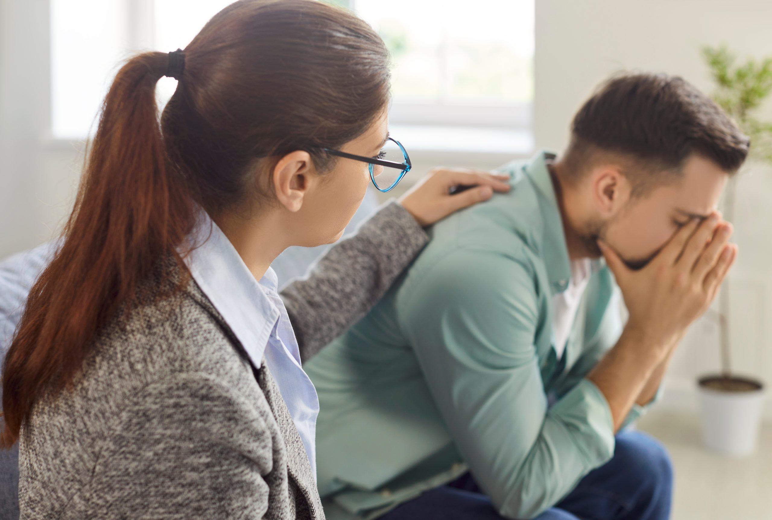 Woman comforting a man from behind while he holds his head in his hands, appearing to be in pain or distress