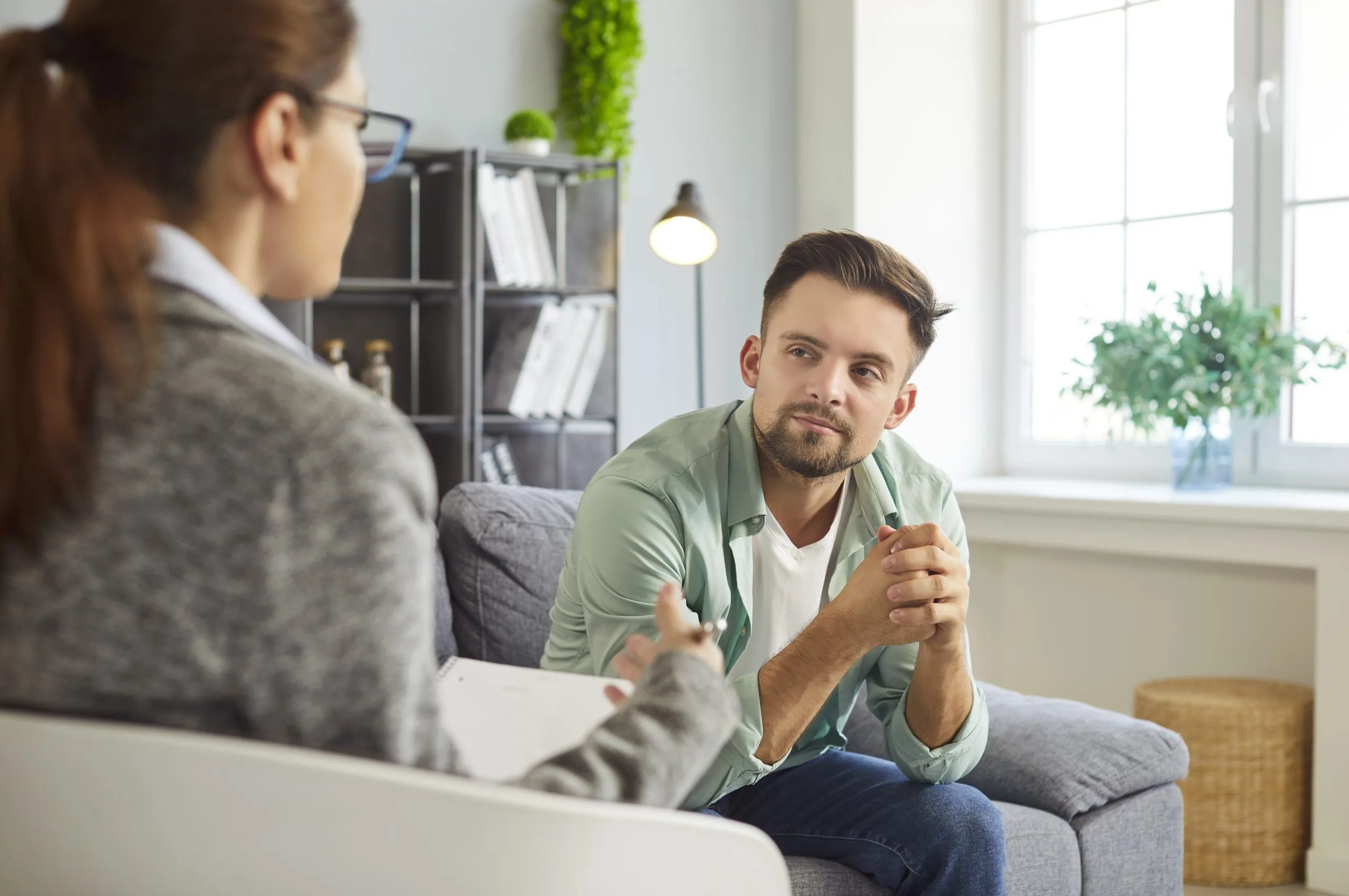 Male therapist gesturing while speaking with female client on couch in bright therapy office