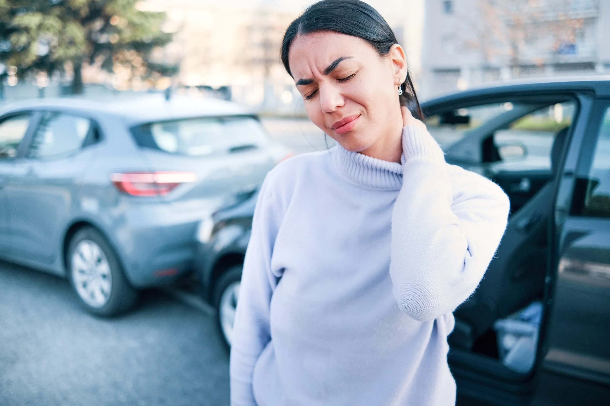 Woman in parking lot wincing while holding her neck, indicating neck pain