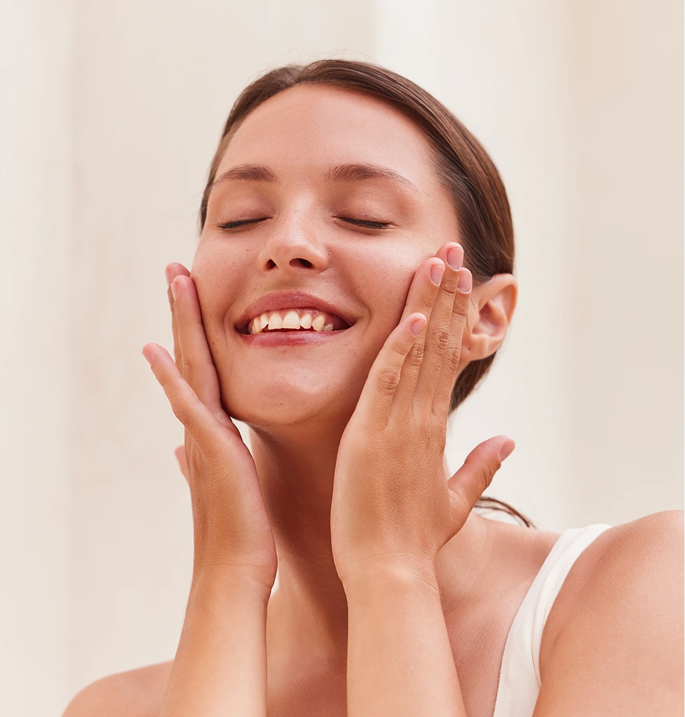 Woman with hands on cheeks smiling with eyes closed against neutral background