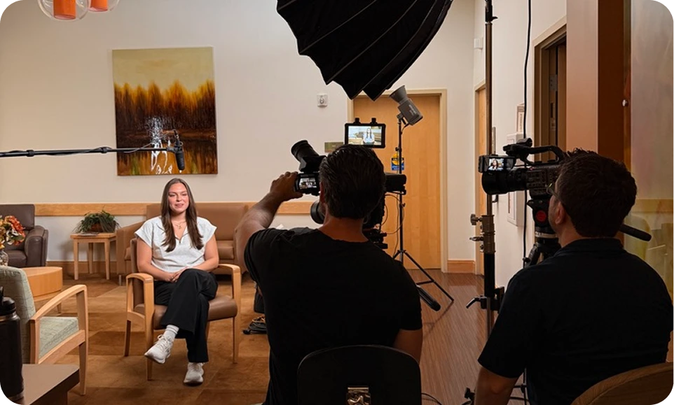 Video production crew filming a woman seated on a chair in a modern office setting with professional lighting and camera equipment