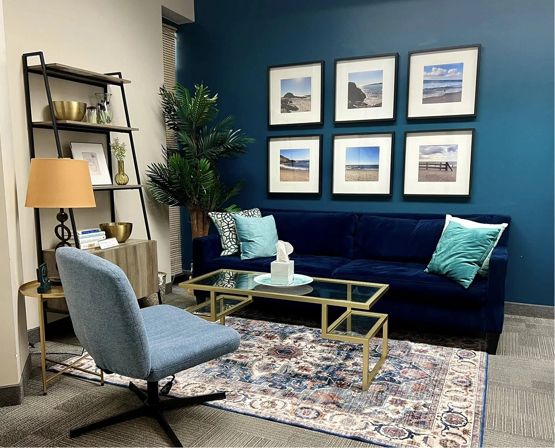 Counseling office waiting room with deep blue sofa, gray accent chair, landscape artwork, and shelving with plants and décor