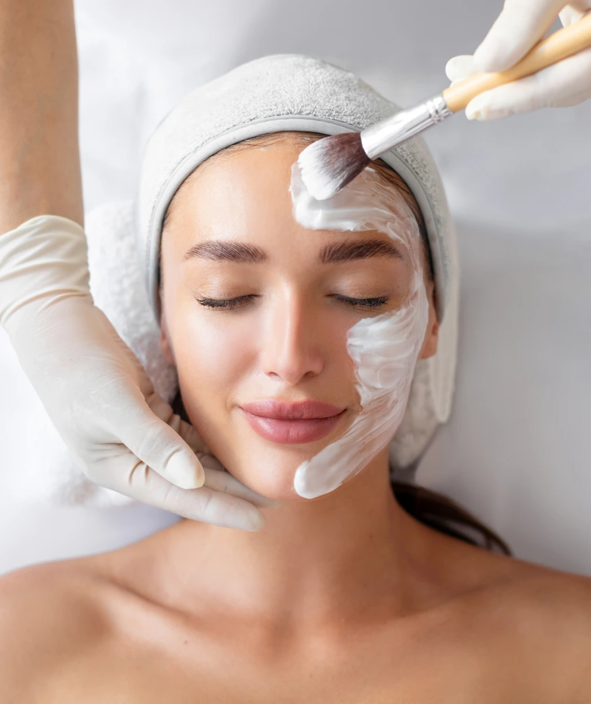 Aesthetician applying white facial mask to patient's forehead with brush during spa treatment