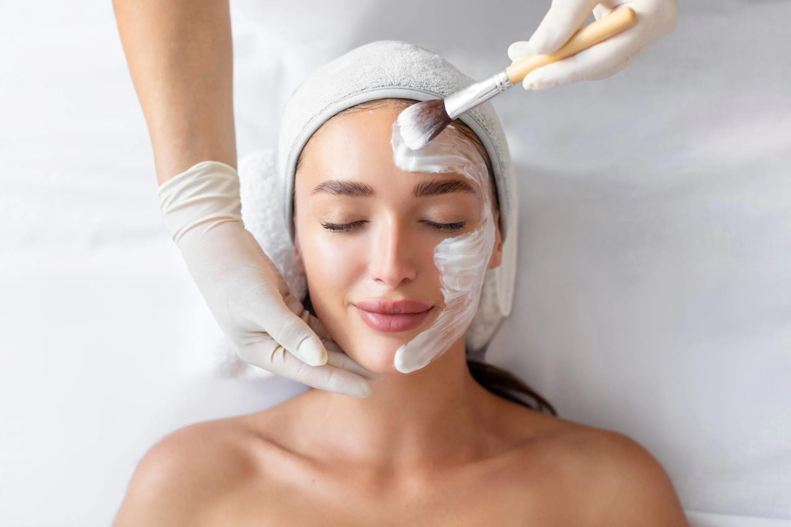Esthetician applying white facial mask to a relaxed woman's face during a professional skincare treatment