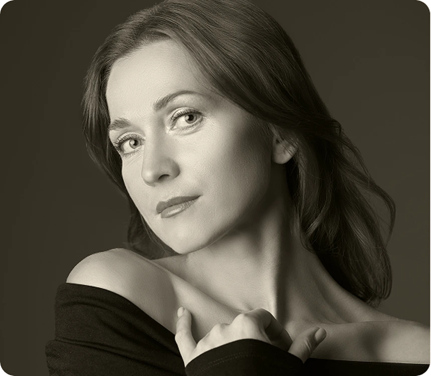 Woman with dark hair wearing black off-shoulder top, looking over shoulder at camera