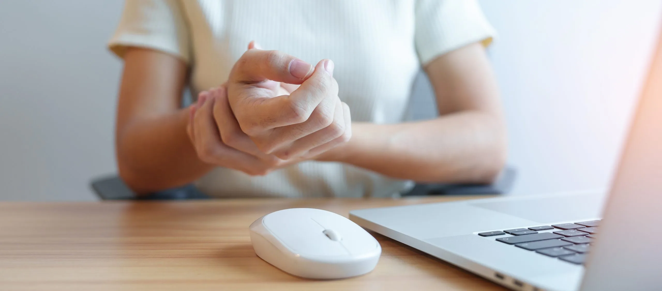 Person holding wrist with visible bandage while seated at desk with laptop and mouse