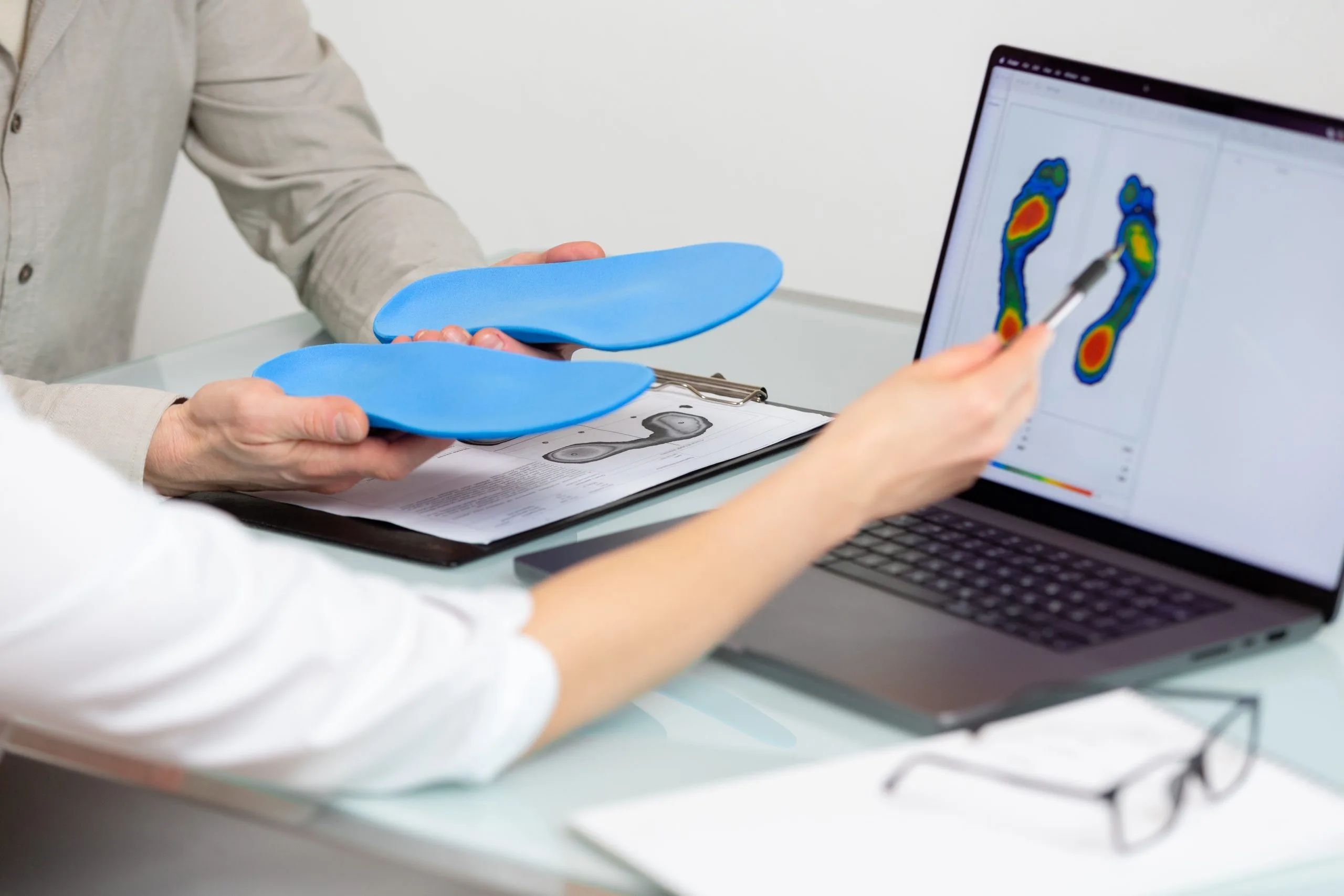 Healthcare provider holding blue orthotic insoles while displaying foot pressure analysis on laptop