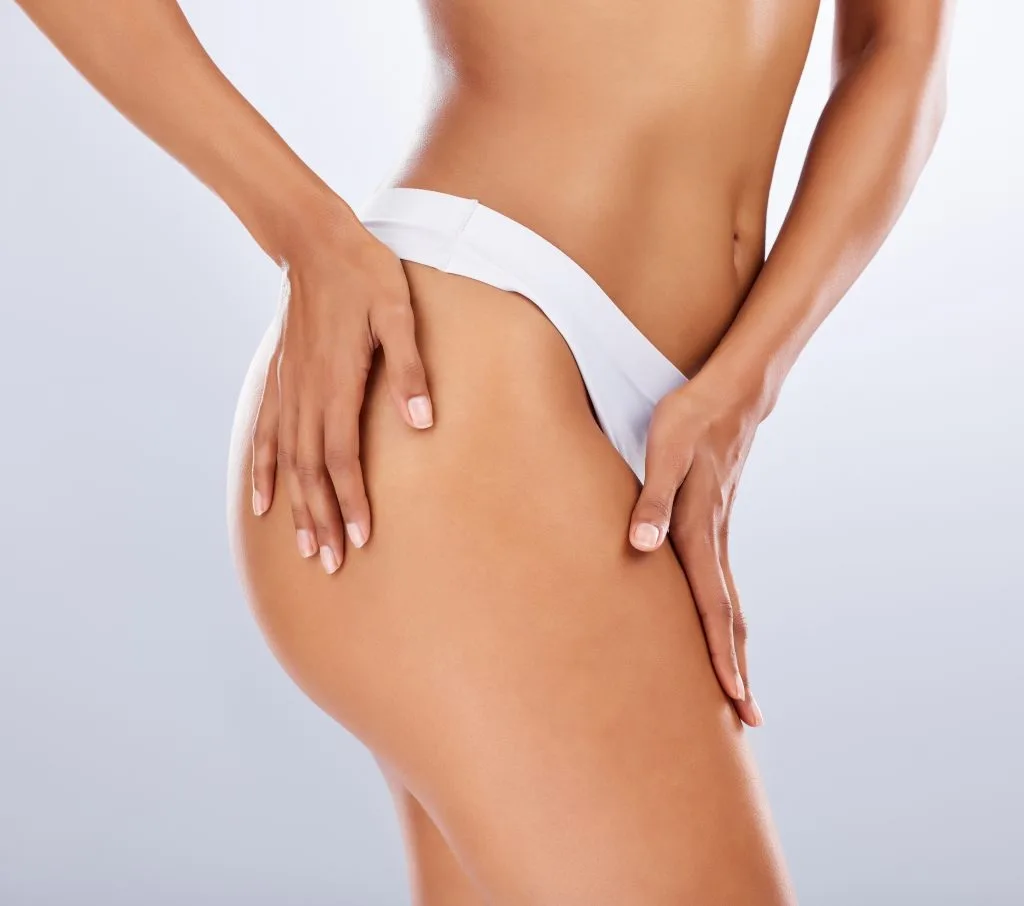 Woman in white bikini bottom examining her thigh and hip contours against a neutral background