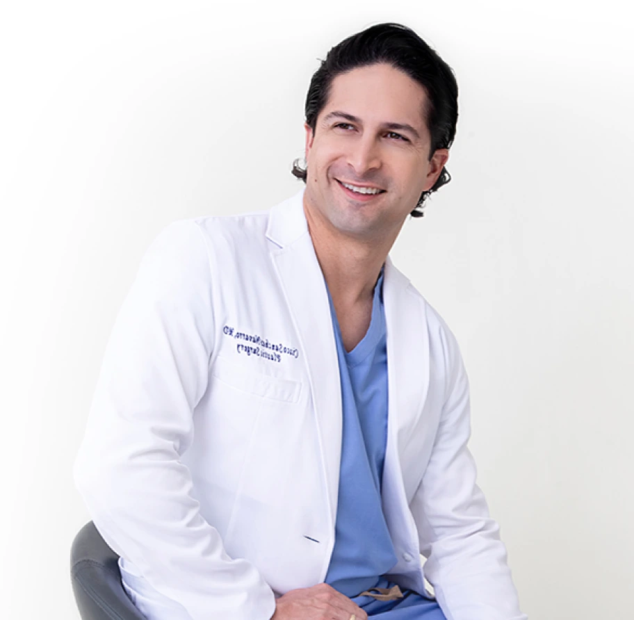 Plastic surgeon in white coat and blue scrubs smiling while seated