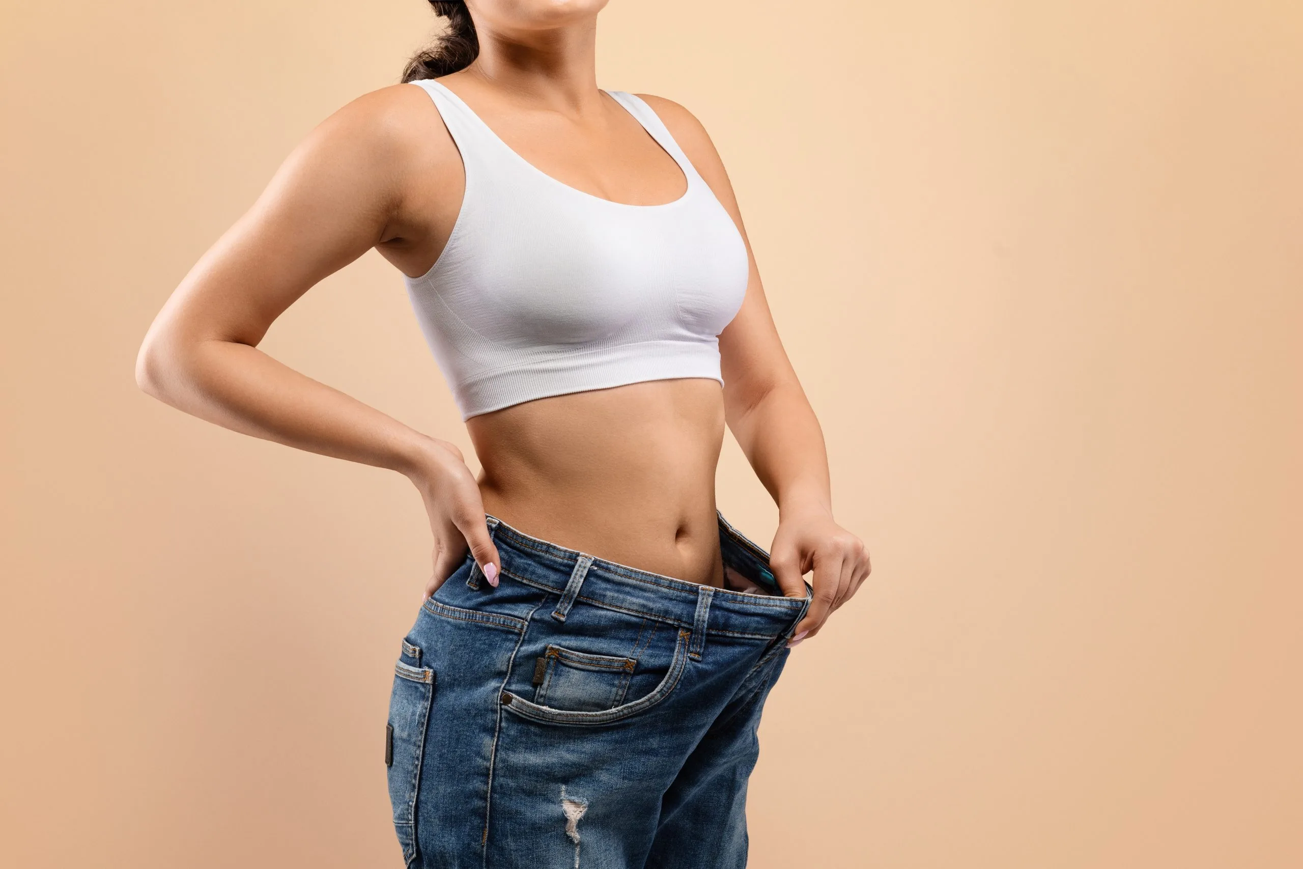 Woman demonstrating weight loss by pulling oversized jeans away from her toned abdomen