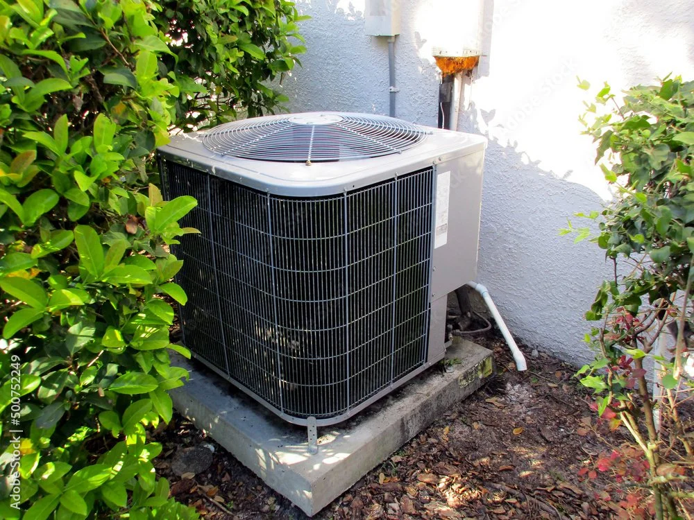 Outdoor AC condenser unit mounted on concrete pad against white house wall surrounded by green landscaping