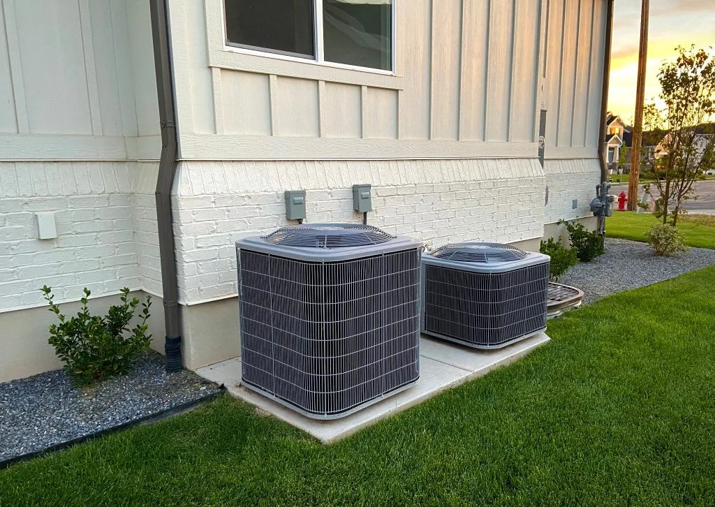 Two residential AC condenser units installed on a concrete pad beside a white brick house with green lawn