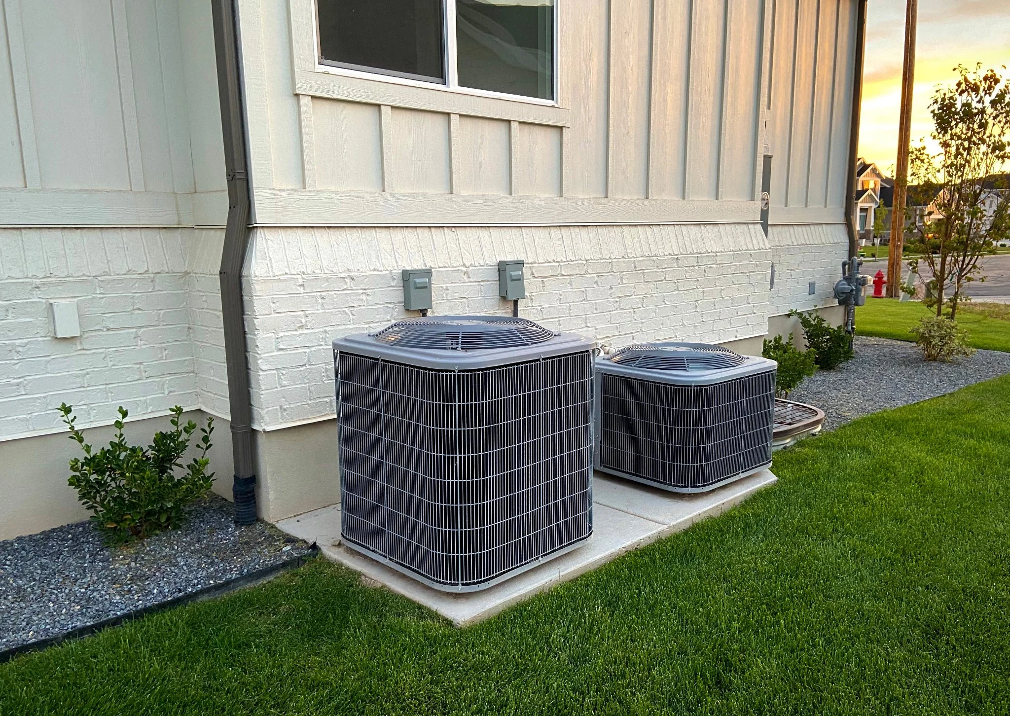 Two residential AC condenser units installed on a concrete pad beside a white brick house with green lawn