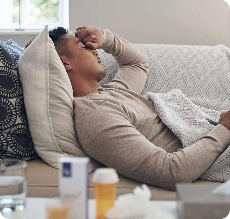 Man resting on a couch with his hand to his face, surrounded by tissues and medications