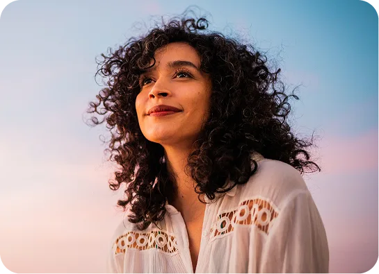 Woman with curly dark hair looking upward against a gradient blue and pink sky