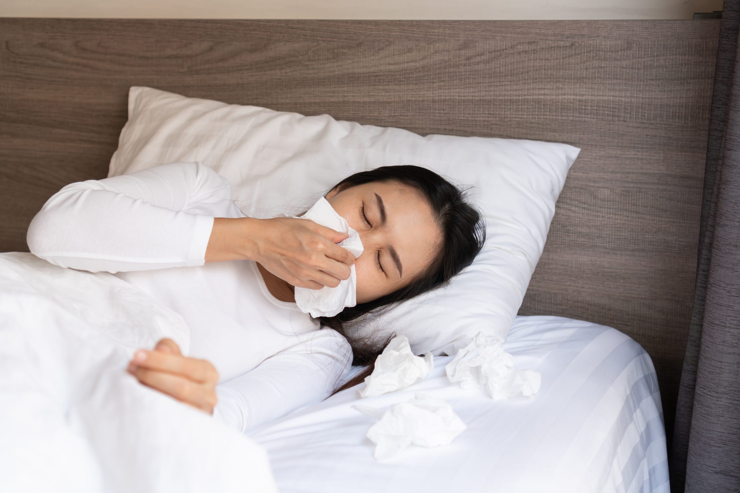Woman in bed blowing her nose into a tissue, showing allergy or flu symptoms