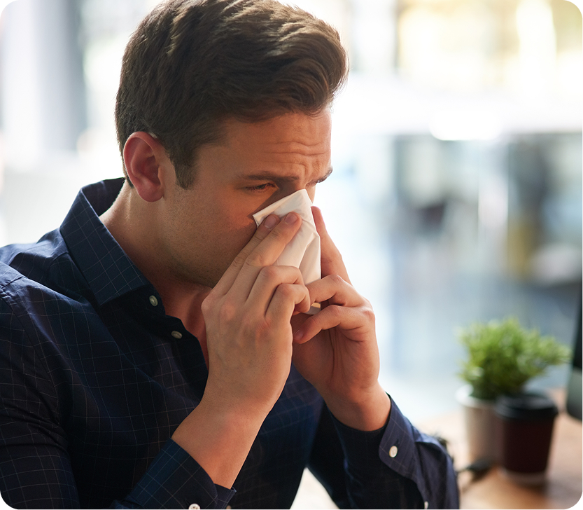 Man in dark blue shirt blowing his nose with tissue at a bright indoor workspace
