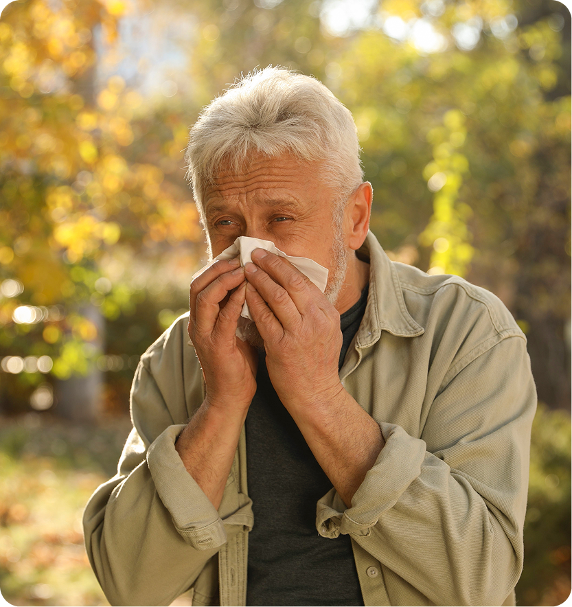 Older man blowing his nose with a tissue outdoors surrounded by yellow fall foliage
