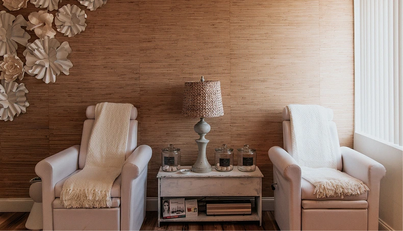 Spa relaxation lounge with two cream armchairs, decorative flowers, lamp, and candles on side table