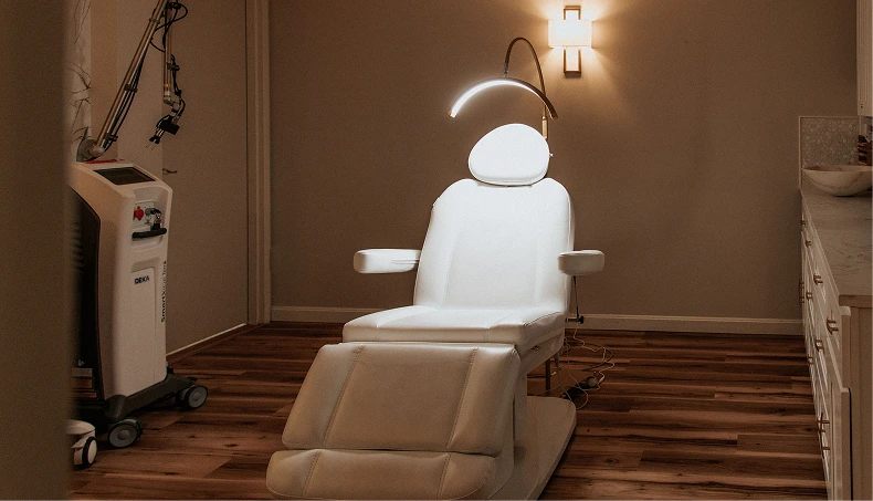 Modern aesthetic treatment room with white exam chair, overhead LED light, and laser equipment