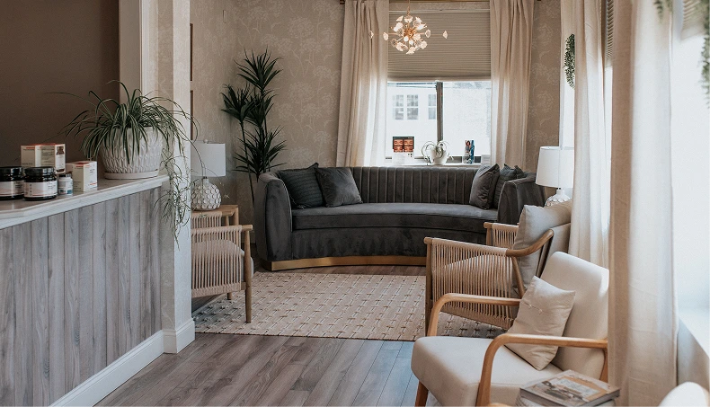 Modern spa waiting area with gray sofa, wooden accent chairs, potted plants, and neutral décor