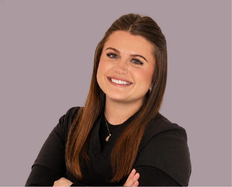 Woman with long brown hair wearing black blazer, arms crossed, smiling at camera