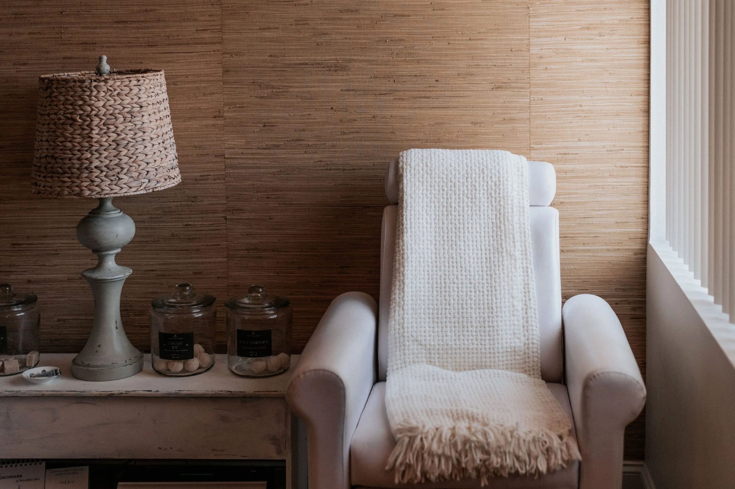 Spa relaxation room with cream upholstered chair draped in white blankets, adjacent side table with decorative lamp and glass candle jars
