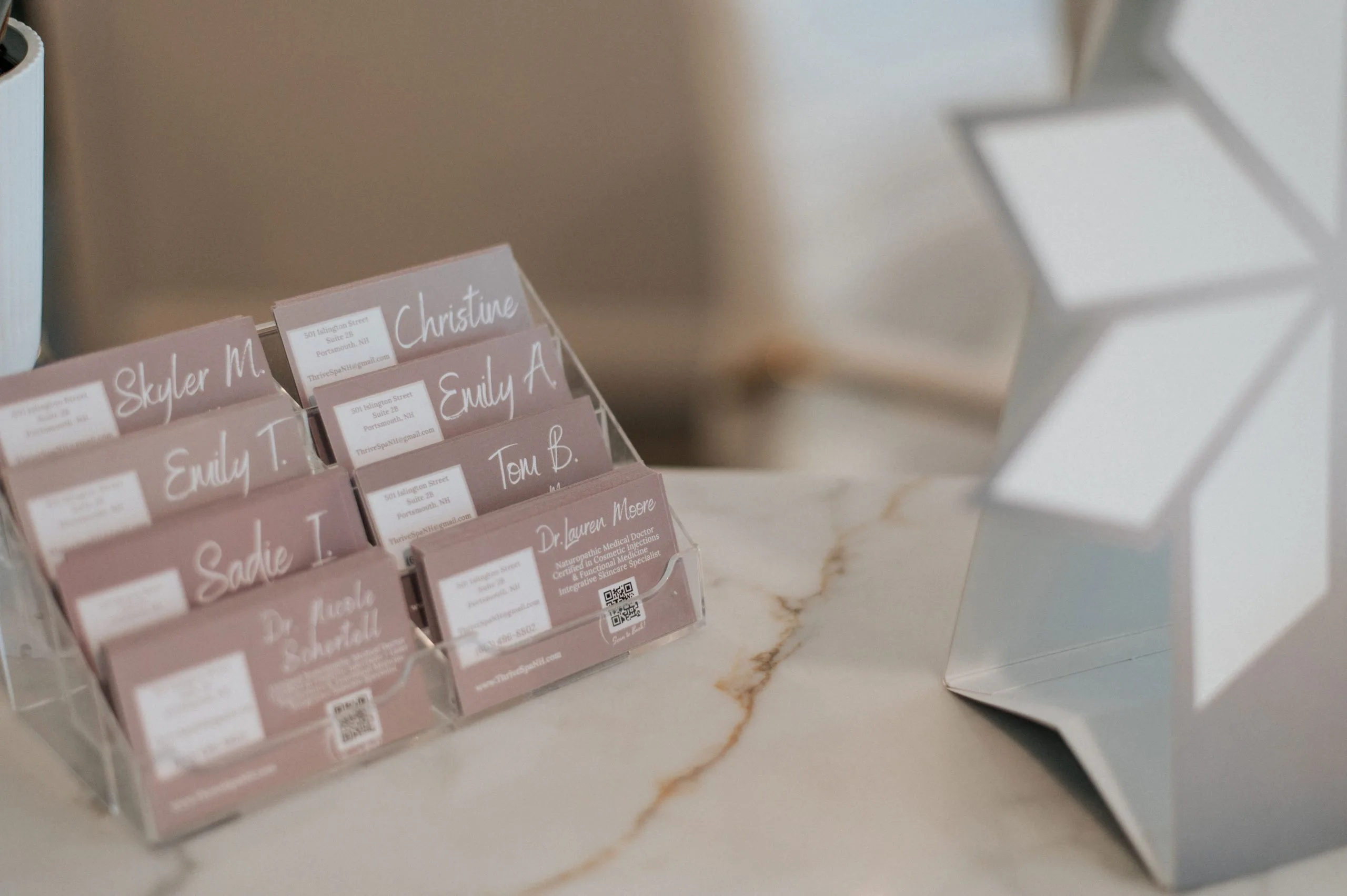 Display of pink business cards for Thrive Med Spa providers including Skyler M., Emily T., Emily A., Christine, Tom B., and Dr. Lauren Moore on a marble reception desk