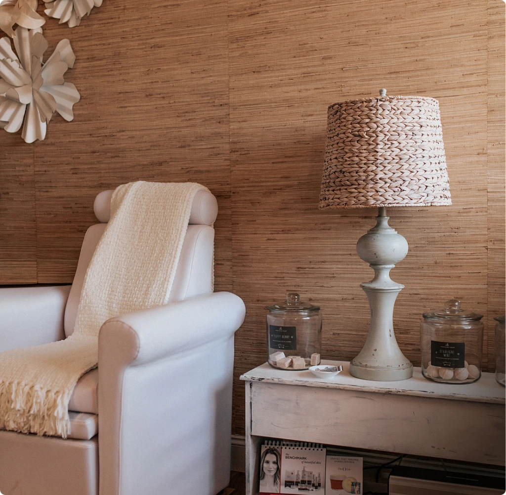 Serene spa room with cream armchair, woven lamp, and decorative candles on whitewashed dresser