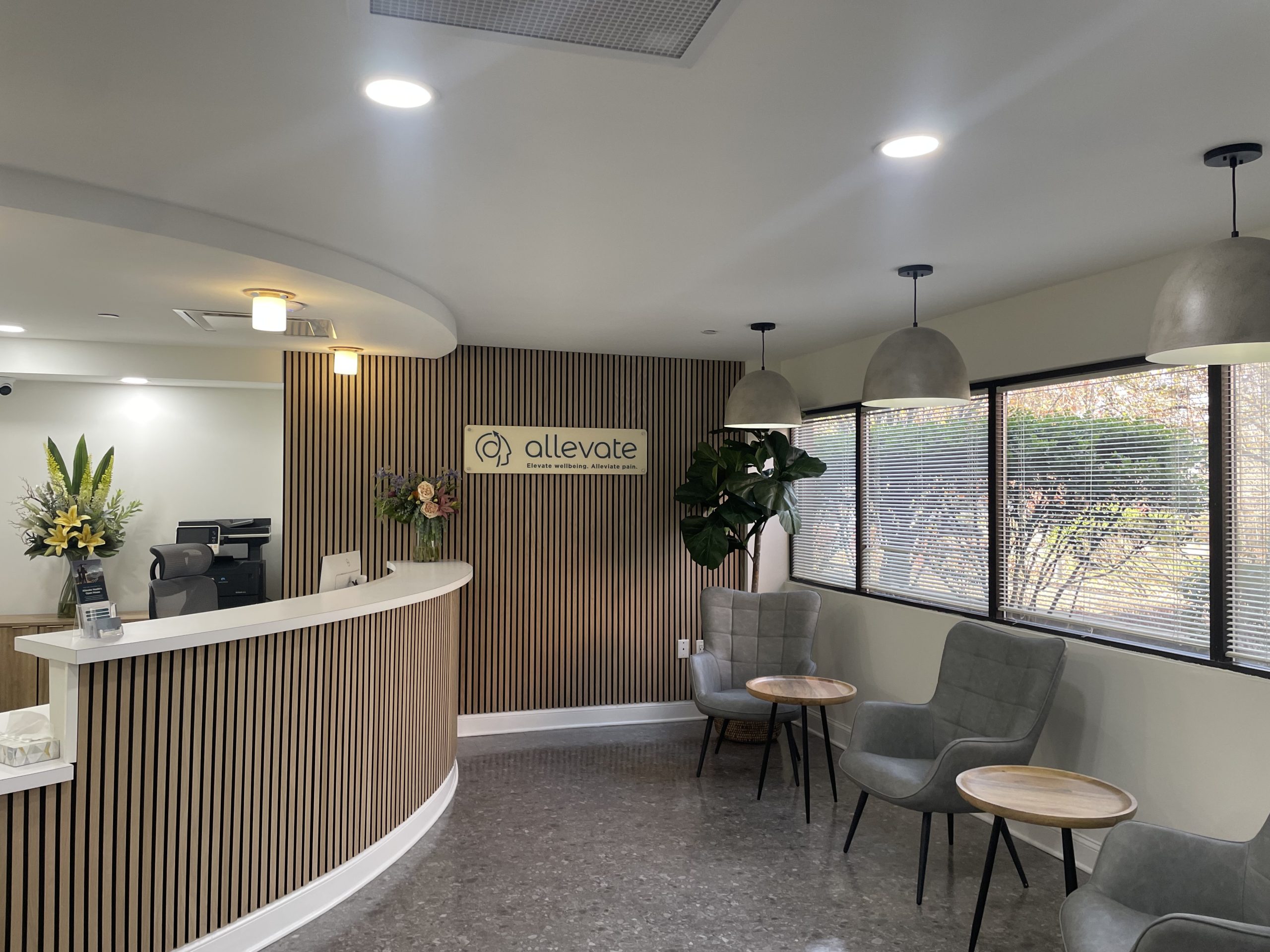 Allevate Health reception area with curved front desk, wood-slat accent wall, logo signage, waiting seating, and potted plants