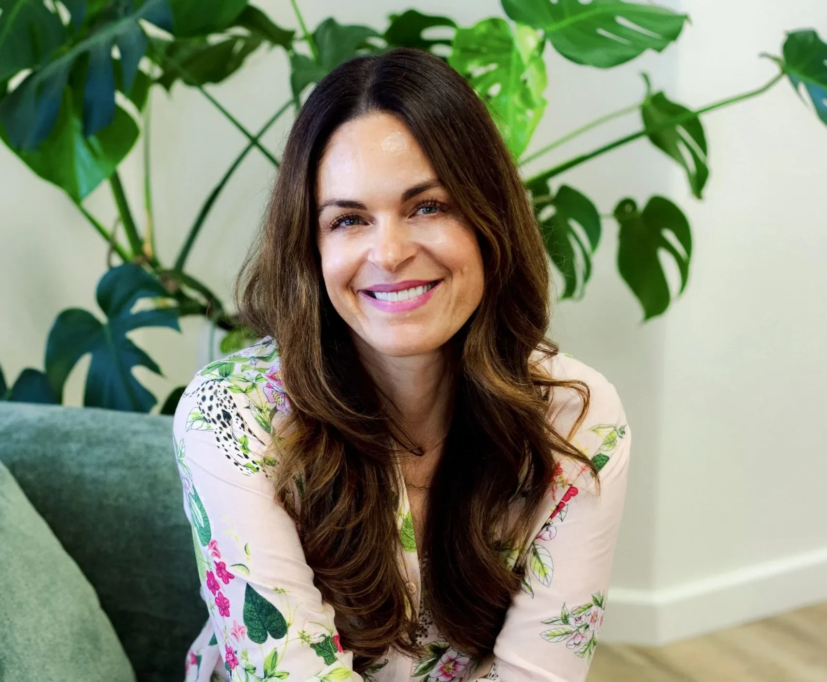 Smiling woman in floral blouse sitting on green chair with potted plant behind her