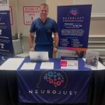Provider standing at Neurojust booth displaying TMS therapy information and equipment at medical event