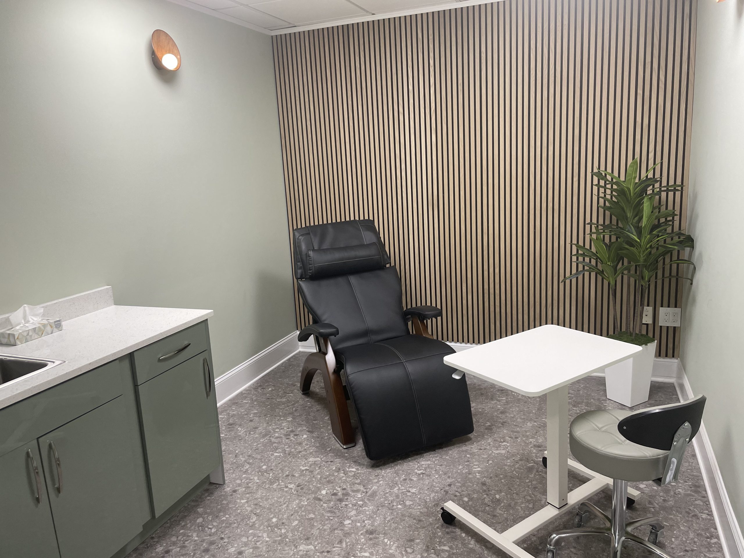 Modern treatment room with black reclining chair, green cabinetry, white tables, and vertical slat wall paneling