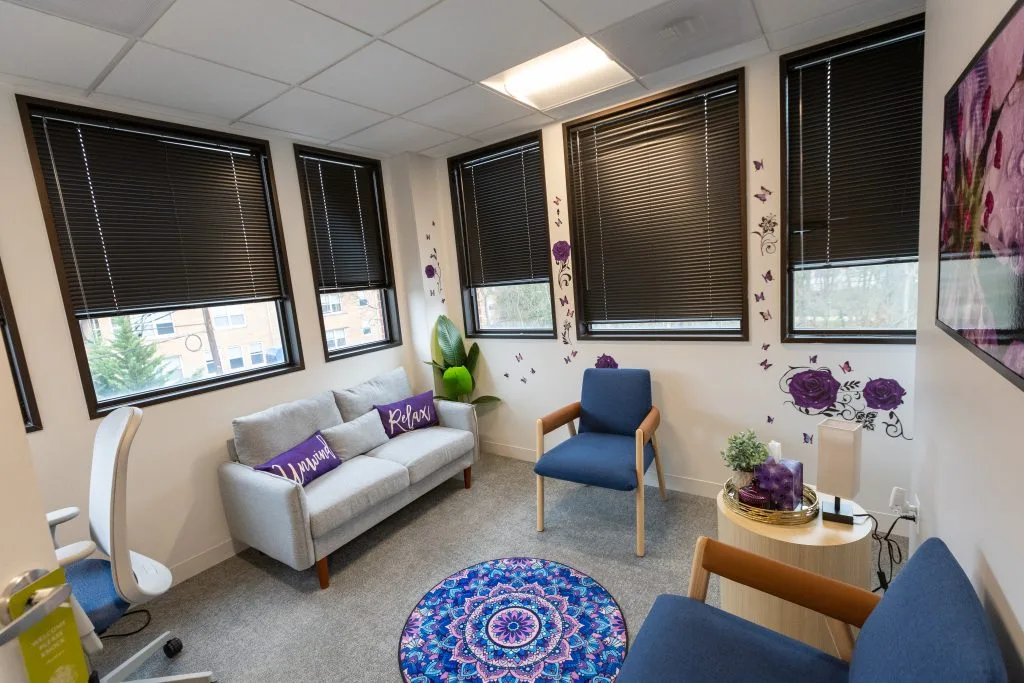Welcoming waiting room with gray sofa, blue chair, decorative purple floral wall decals, and mandala-patterned floor rug