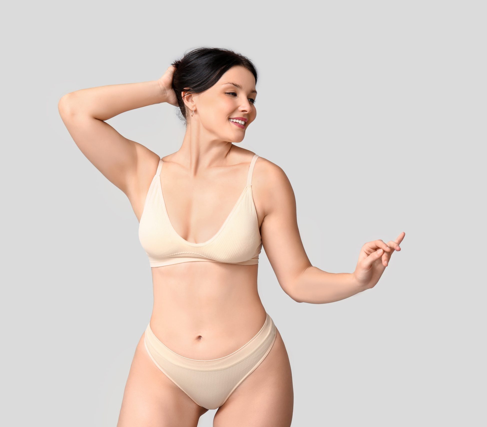 Woman in neutral-toned bikini with hand behind head smiling against light gray background