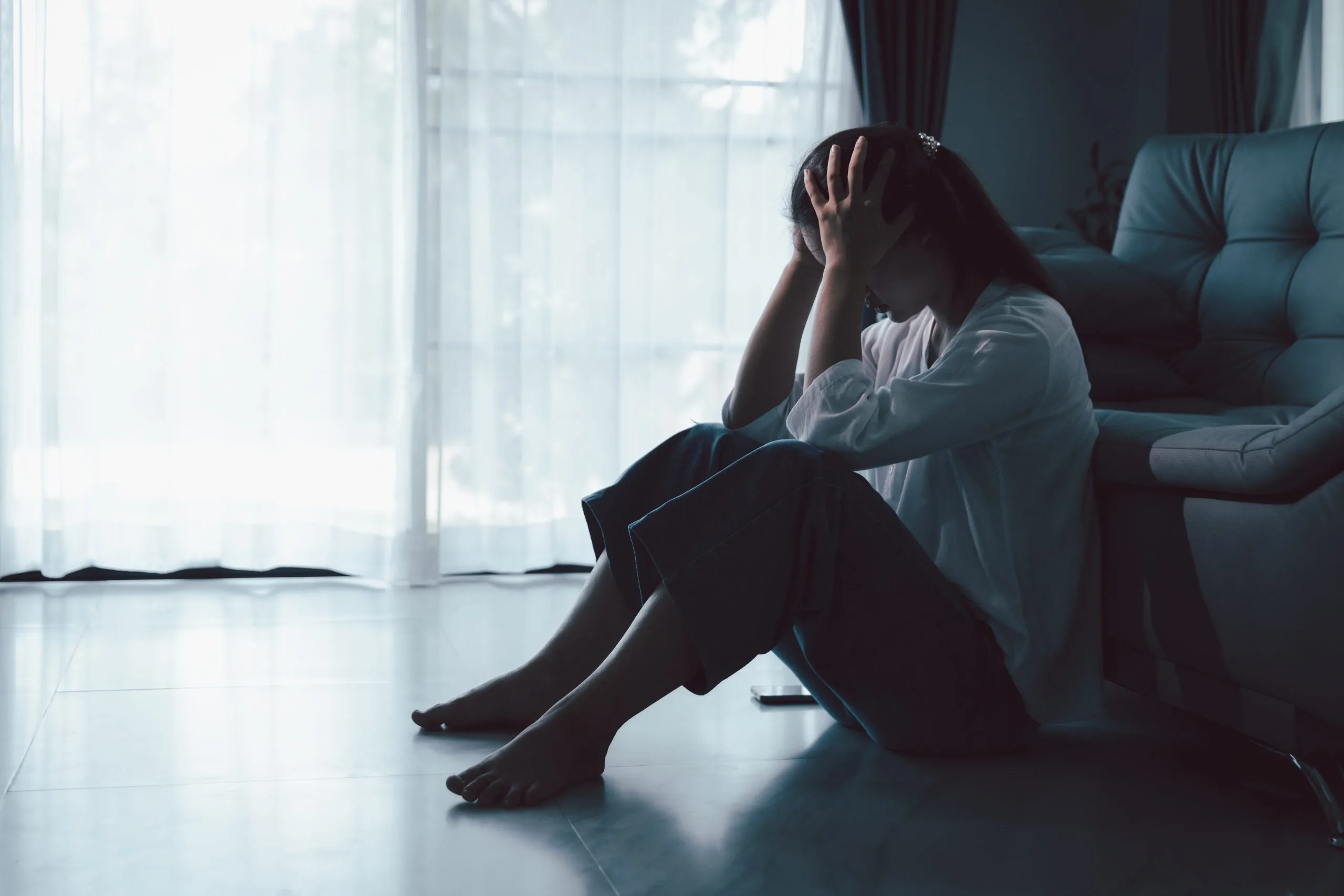 Woman sitting on floor against couch with head in hands, looking distressed by a window